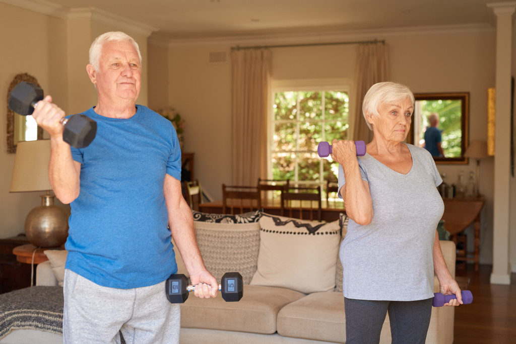 Senior adult man and woman exercising using weights in the living room of their home.