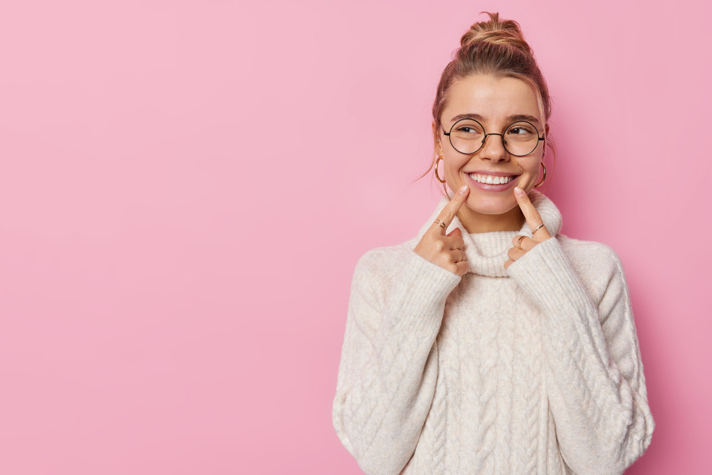 young woman with glasses smiling pointing to her teeth against pink background