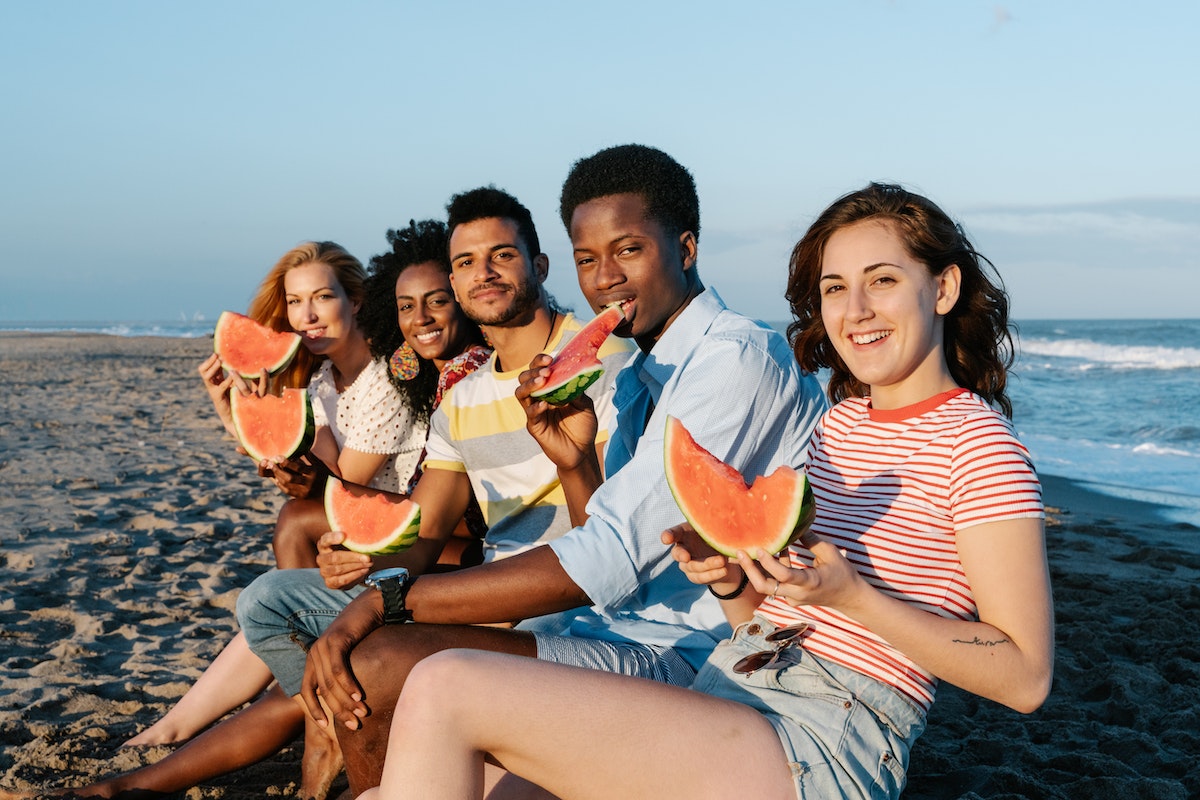 Glad diverse travelers eating watermelon on ocean beach